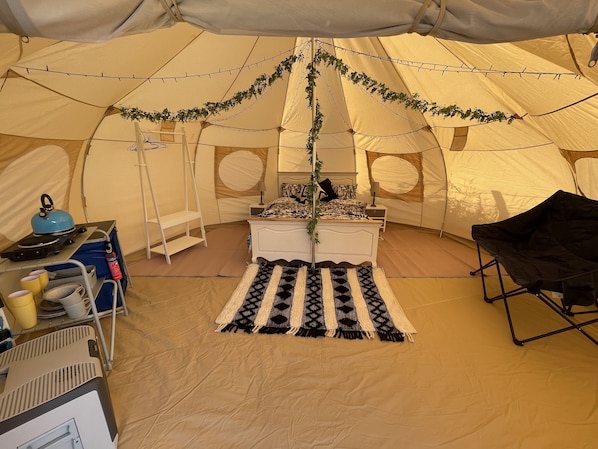 A tent with a bed, a small table, a chair, and a shelf with items.