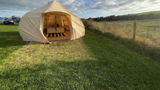 A large, beige tent with a bed and a small table inside, set up on a grassy field with a fence in the background. - Impeccable 1-bed Bell Tent Near Holyhead (Holyhead)