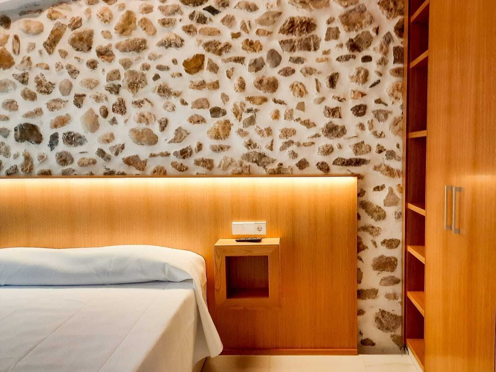 Un dormitorio con pared de piedra decorativa, cabecero de madera, mesita de noche con lámpara y estanterías empotradas de madera.