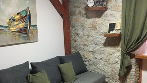 A cozy living room with a grey sofa, green cushions, a painting of a sailboat, a clock, and a bicycle on a stone wall. - Apartment Equinox-Island Pag (Pag)