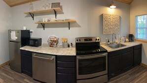 A modern kitchen with a built-in oven, microwave, and dishwasher, wooden shelves above the counter, and a decorative wall piece. - Cana Cottage | Farm Stay (Tyler)