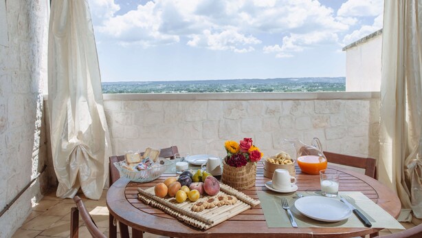 A breakfast table set on a balcony with a view of a distant landscape, a basket of flowers, and various breakfast items.