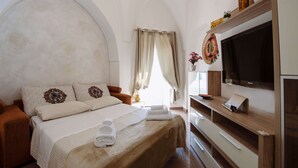 A hotel room with a bed, a TV, a wooden shelf, and a large arched window.