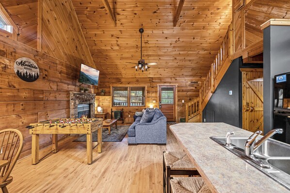 Relax in a cozy cabin-style space with a central seating area, foosball table, and TV for entertainment, plus a kitchen with sink and refrigerator for self-catering.