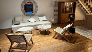 A living room with a sofa, two round tables, and two wicker chairs. A large mirror hangs on the wall, and a staircase is visible in the background. - Charming town with panoramic views and swimming pool (Saint-Leu)