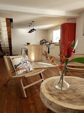 Un soggiorno moderno con pavimento in legno, un tavolino rotondo con un vaso di fiori, una poltrona in vimini con cuscino e una cucina sullo sfondo.