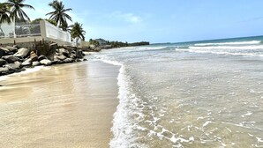 A beach with clear waters, palm trees, and a building with a balcony. - Guest Favorite • Beachfront Luquillo Condo • Walk to Kioskos & El Yunque (Luquillo)