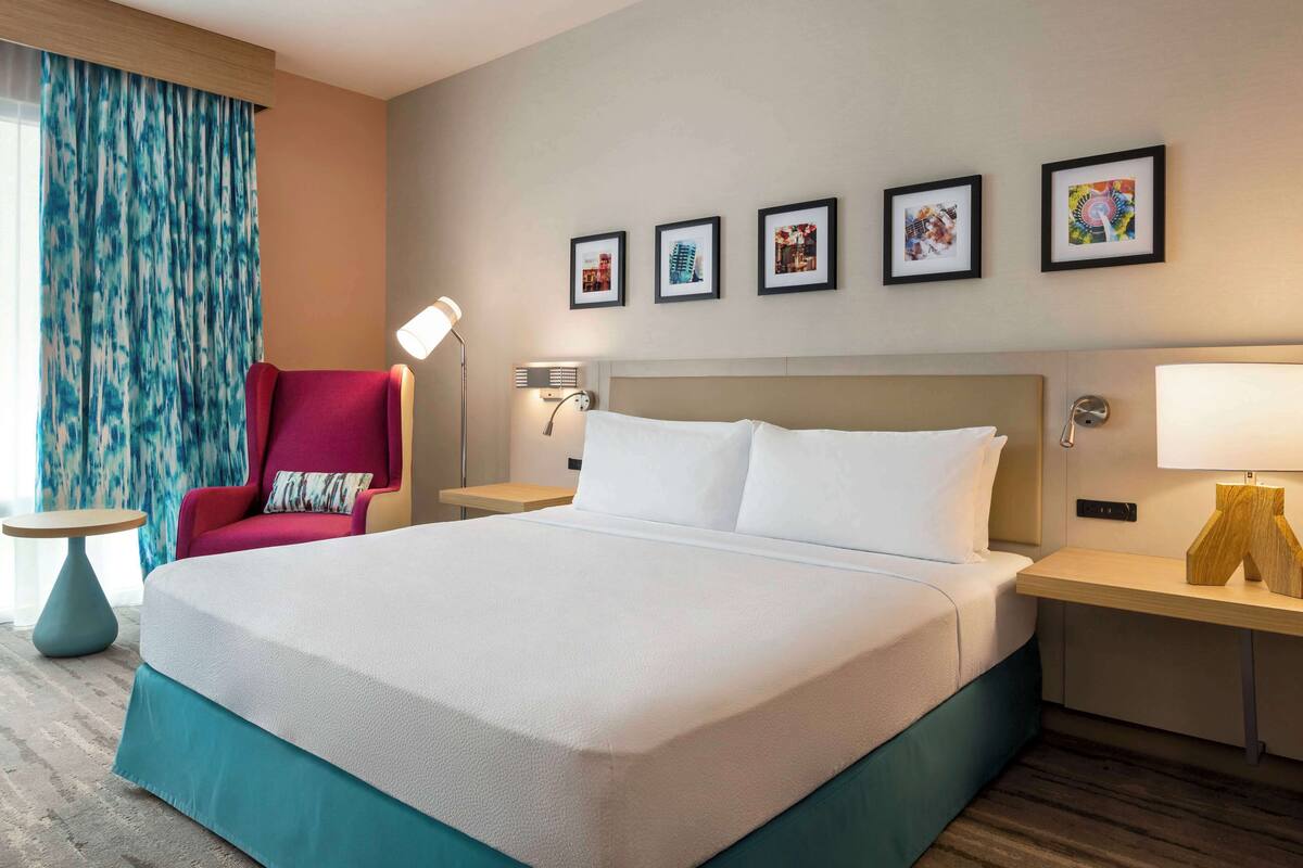 Ein Hotelzimmer mit einem großen Bett, zwei Nachttischen, einem roten Sessel, einer Stehlampe und vier gerahmten Bildern an der Wand.