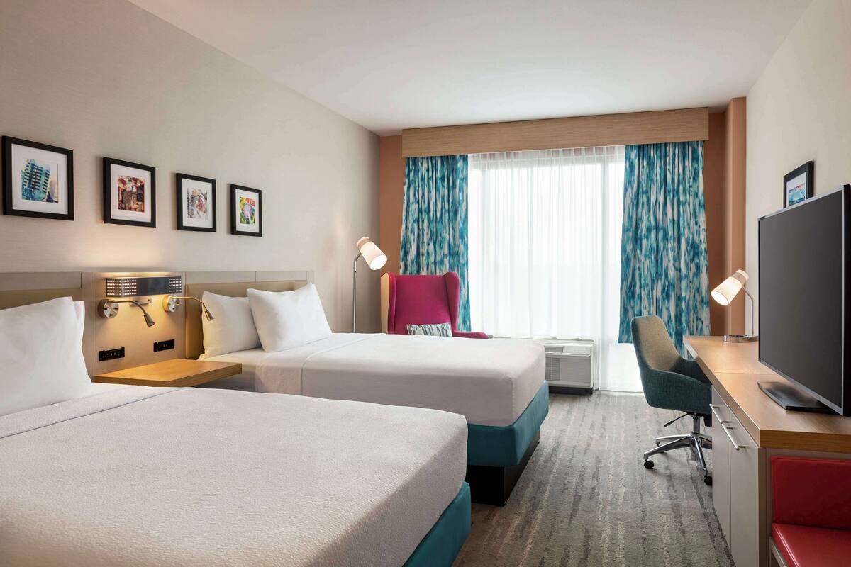 Ein Hotelzimmer mit zwei Betten, einem Schreibtisch, einem Fernseher, einem Sessel und einem Fenster mit Vorhängen.