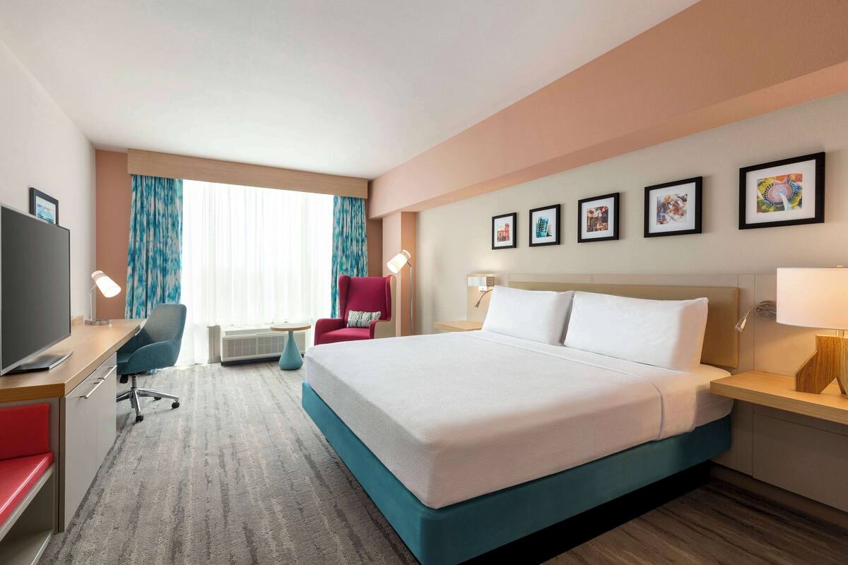 Ein Hotelzimmer mit einem großen Bett, einem Schreibtisch mit Fernseher, einem roten Sessel und einem Fenster mit Vorhängen.