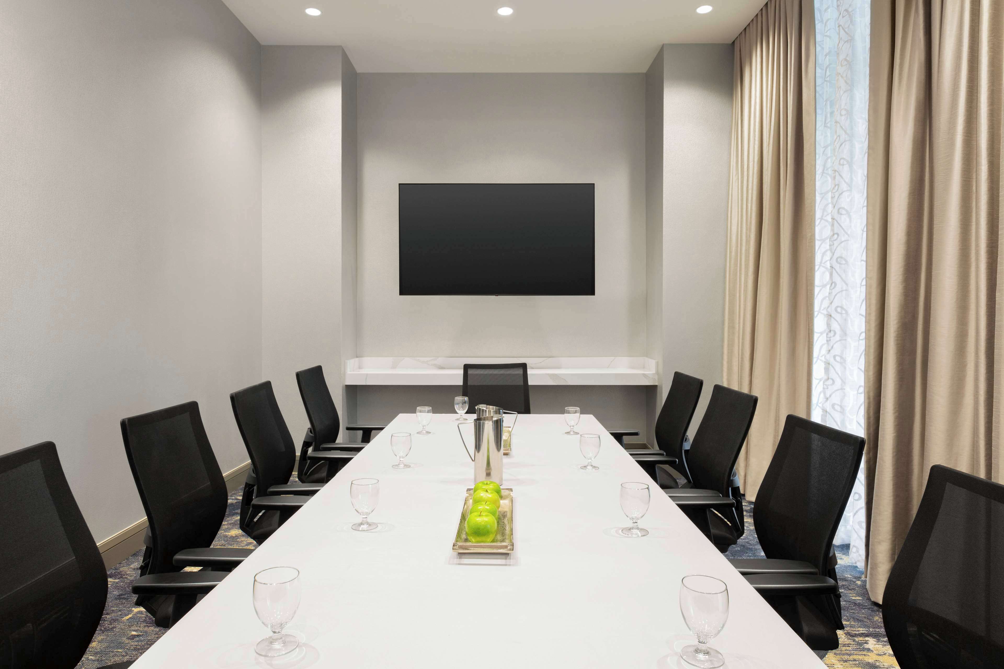 Une salle de conférence avec une longue table, des chaises noires, un écran de projection et une fenêtre avec des rideaux.