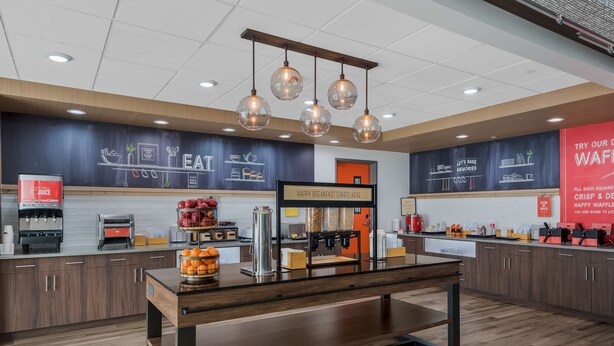 A modern breakfast area with a coffee station, fruit display, and a sign indicating breakfast service. - Hampton Inn Williams (Williams)