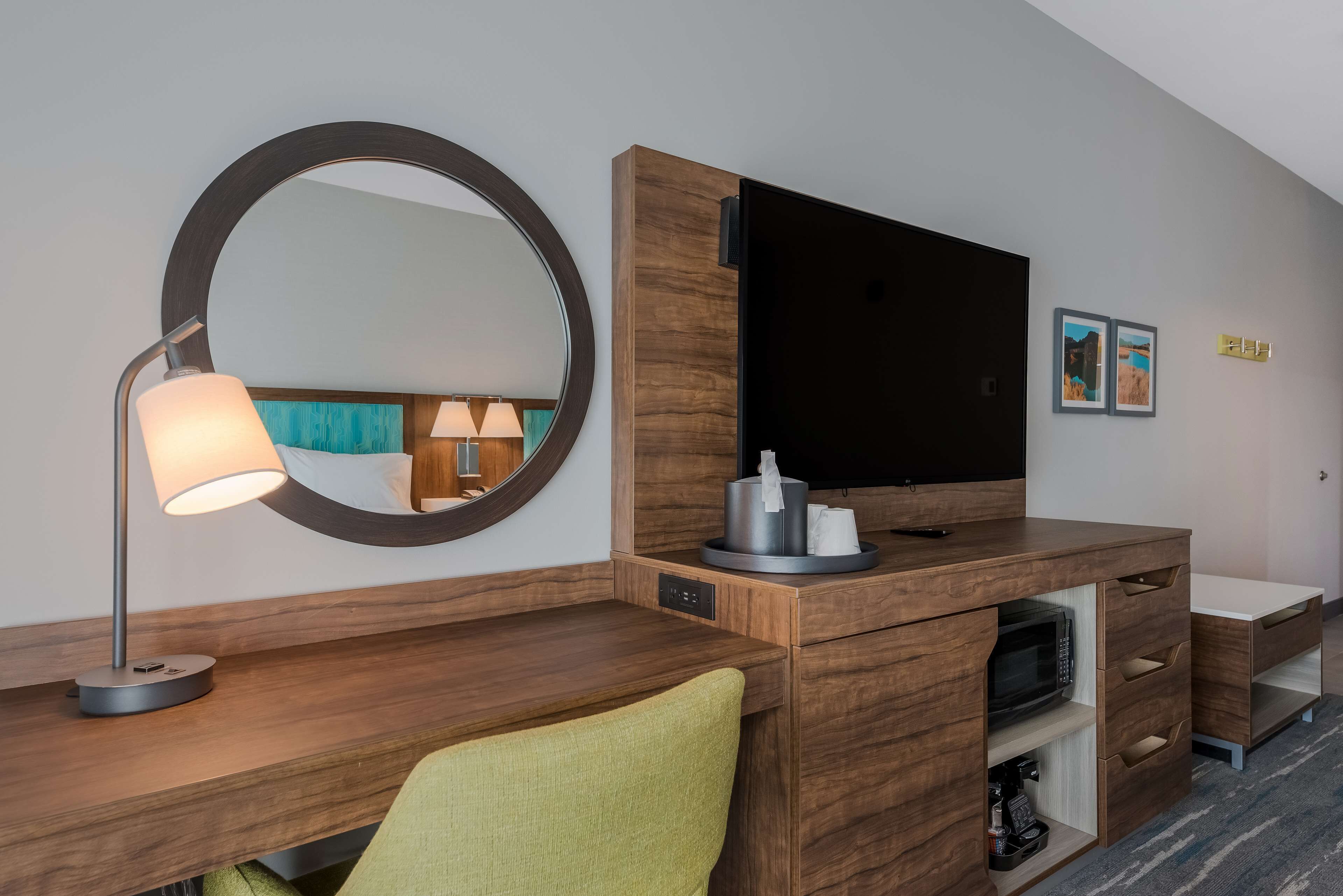 Habitación de hotel con un escritorio de madera, un espejo redondo, un televisor de pantalla plana, una lámpara, un microondas y un pequeño estante con artículos de tocador.