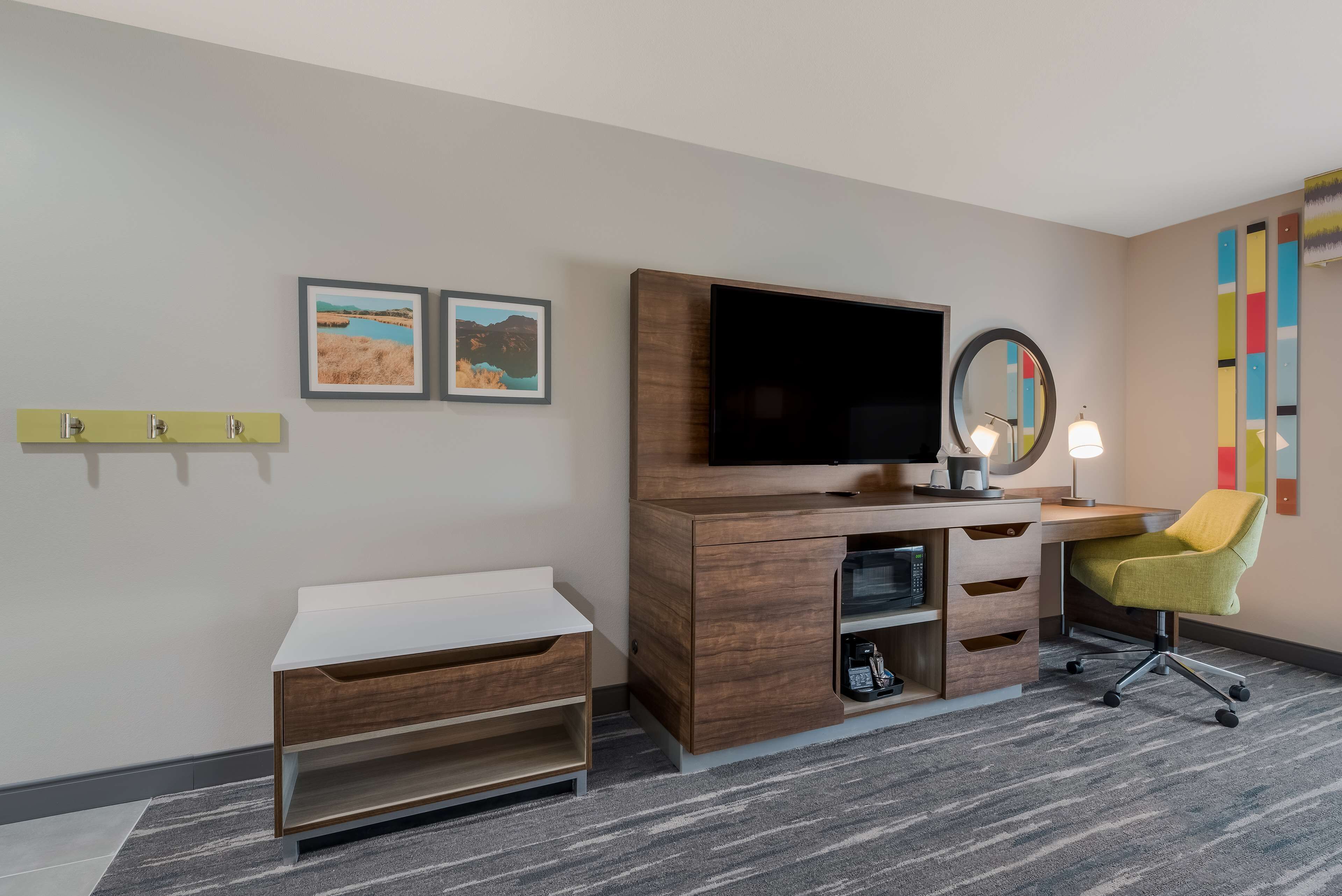 A hotel room with a wooden desk, a flat-screen TV, a small shelf, a chair, and two framed pictures on the wall.