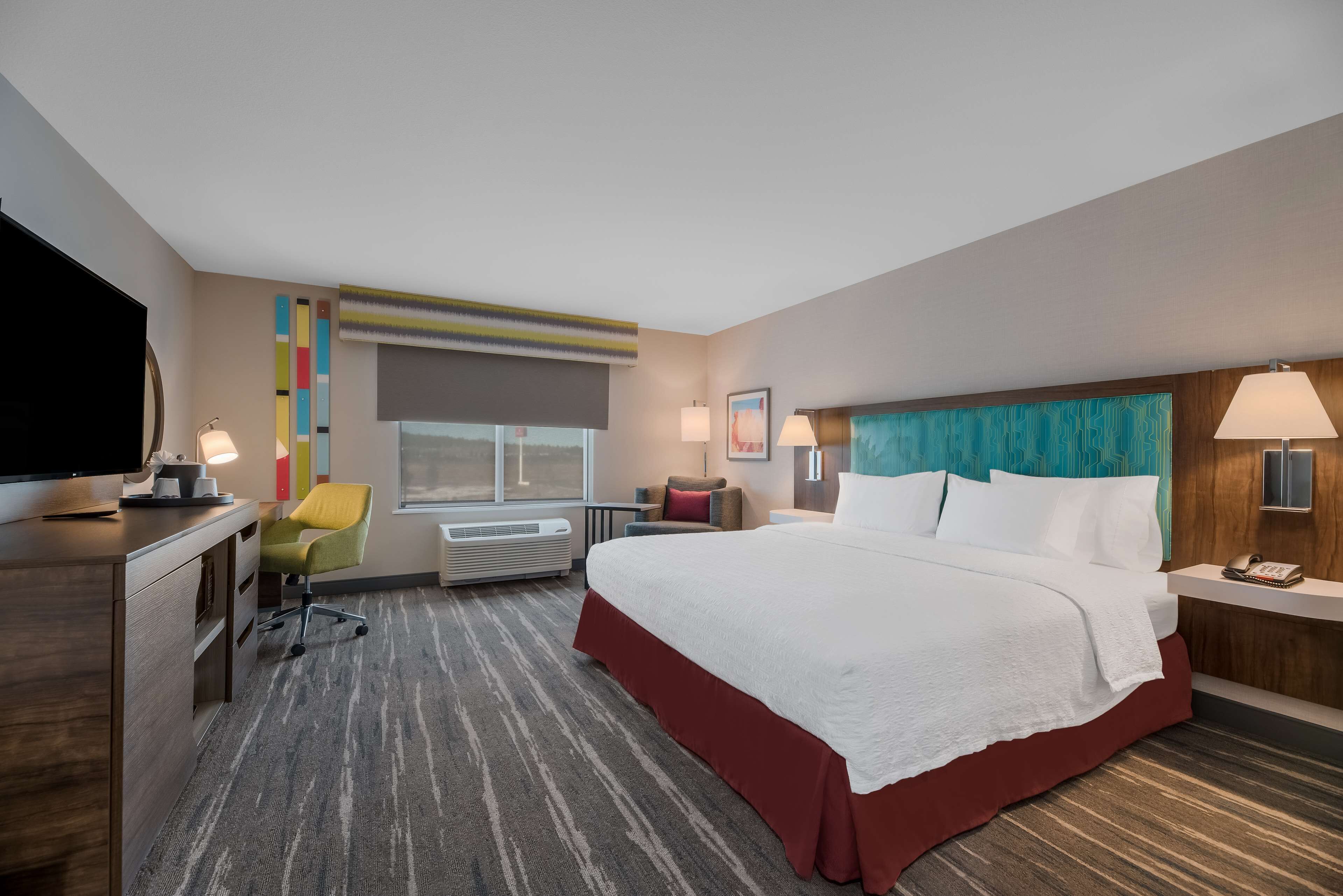 A hotel room with a large bed, a desk with a television, a chair, and a window with blinds.
