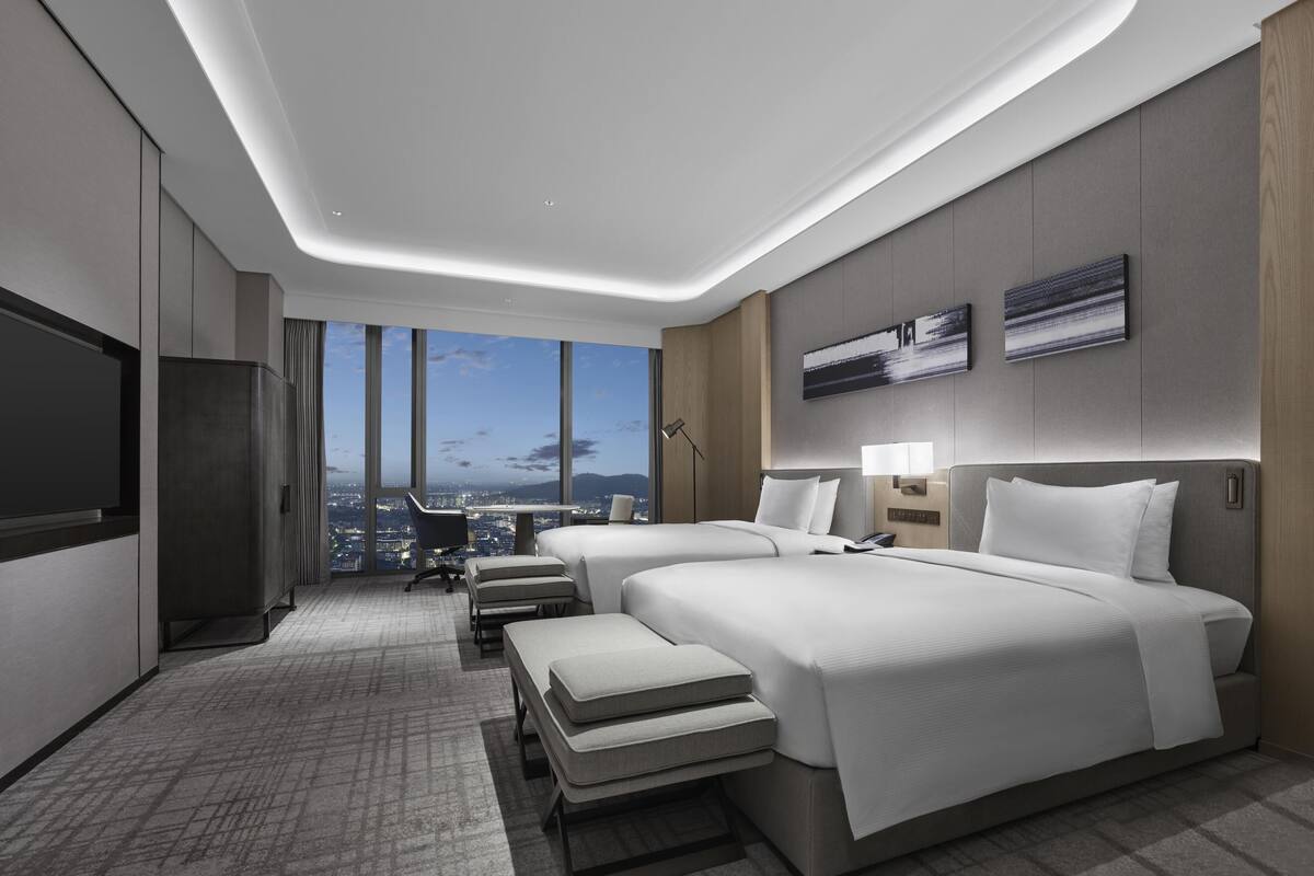 A modern hotel room with two beds, a desk, a chair, a TV, and a city view.