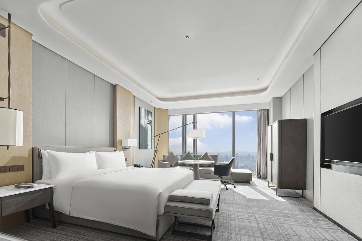 A modern hotel room with a large bed, a desk, a chair, a TV, and a city view.