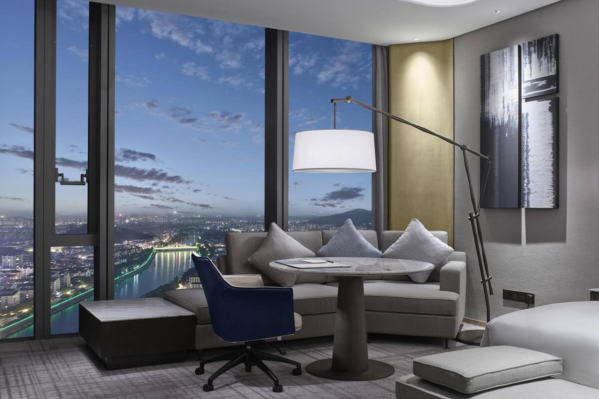 A modern hotel room with a large window, a sofa, a round table, and a floor lamp.