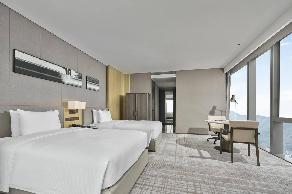 A modern hotel room with two beds, a desk, and a large window offering a city view.