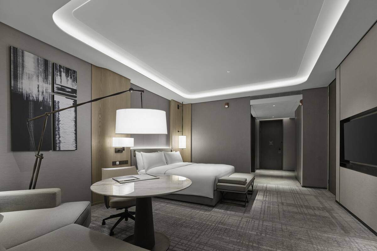 A modern hotel room with a bed, bedside tables, a desk, a chair, a lamp, and artwork on the walls.