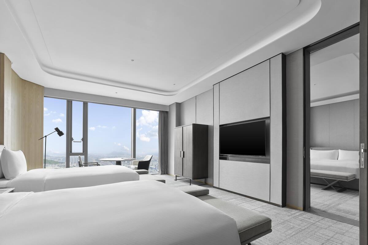 A modern hotel room with two beds, a TV, a desk with a chair, and a large window offering a city view.