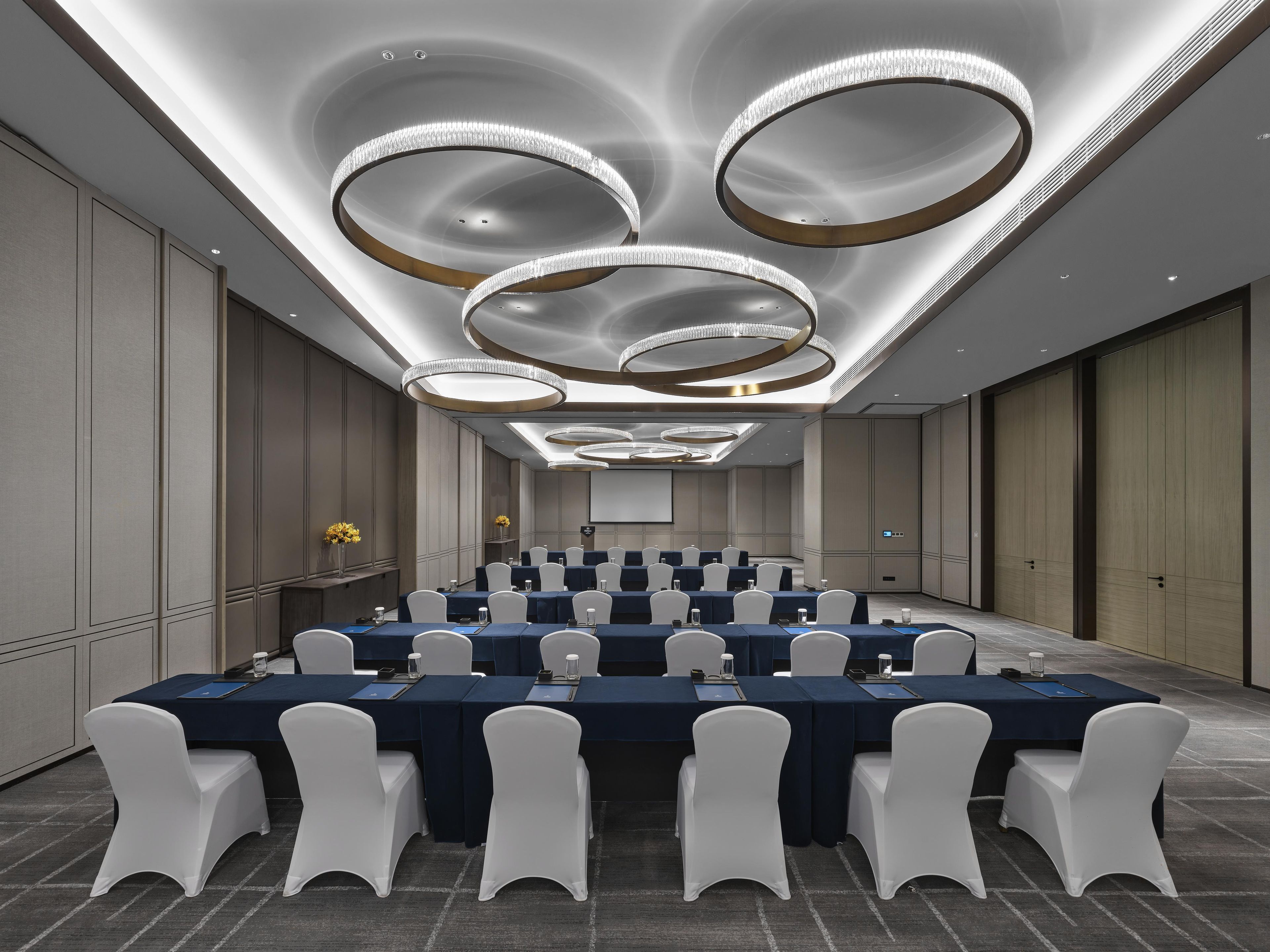 Une salle de conférence dotée de plusieurs tables, de chaises blanches et d’un design moderne au niveau du plafond.