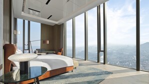 A modern hotel room with a large bed, a glass table, a chair, a lamp, and a view of the city. - Hilton Zhuji (Shaoxing)