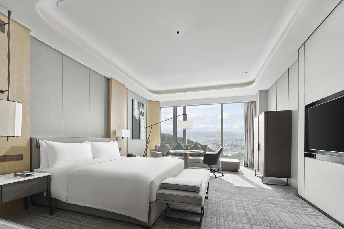 A modern hotel room with a large bed, a desk, a chair, a TV, and a city view.