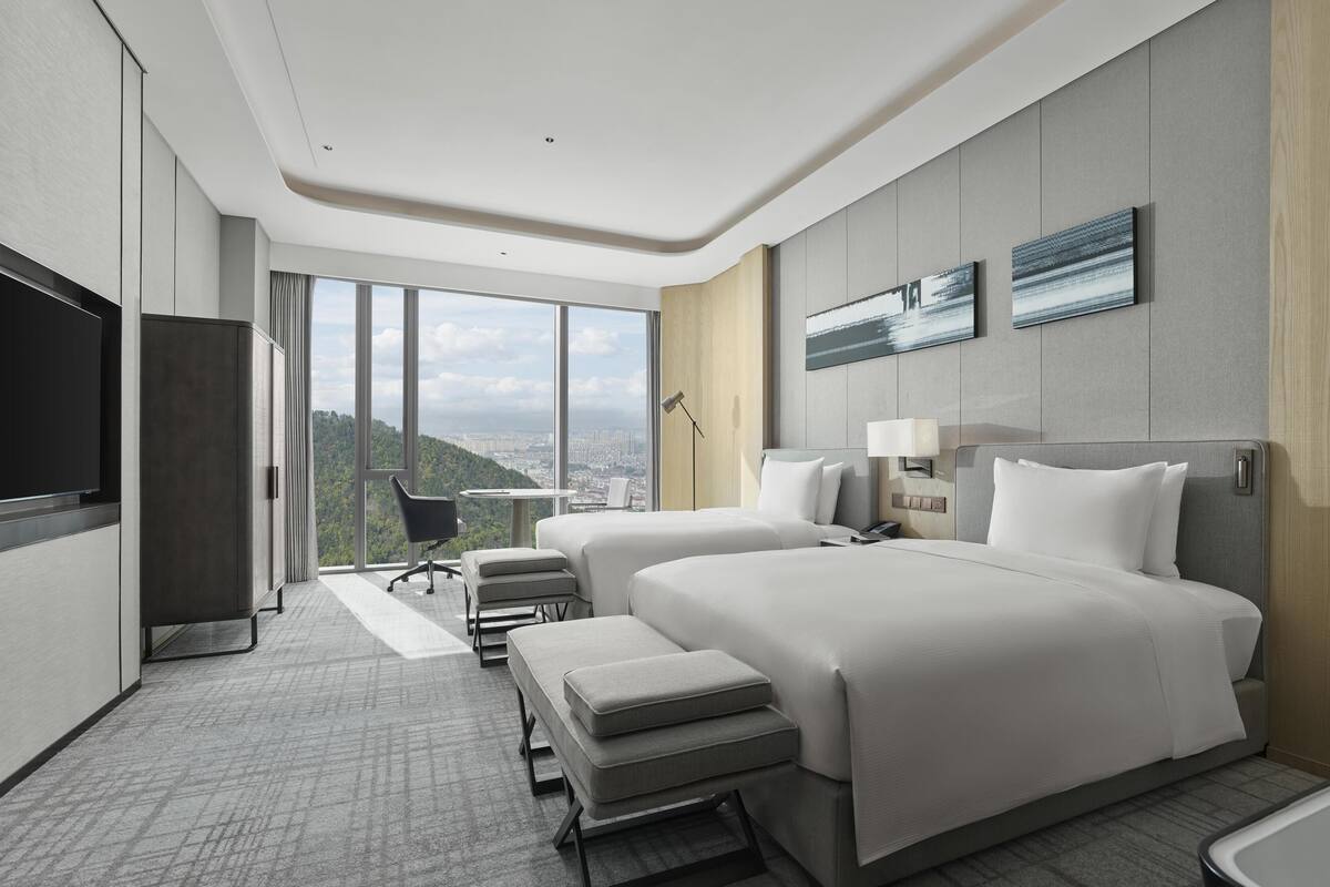 A modern hotel room with two beds, a desk, a chair, and a large window offering a city view.