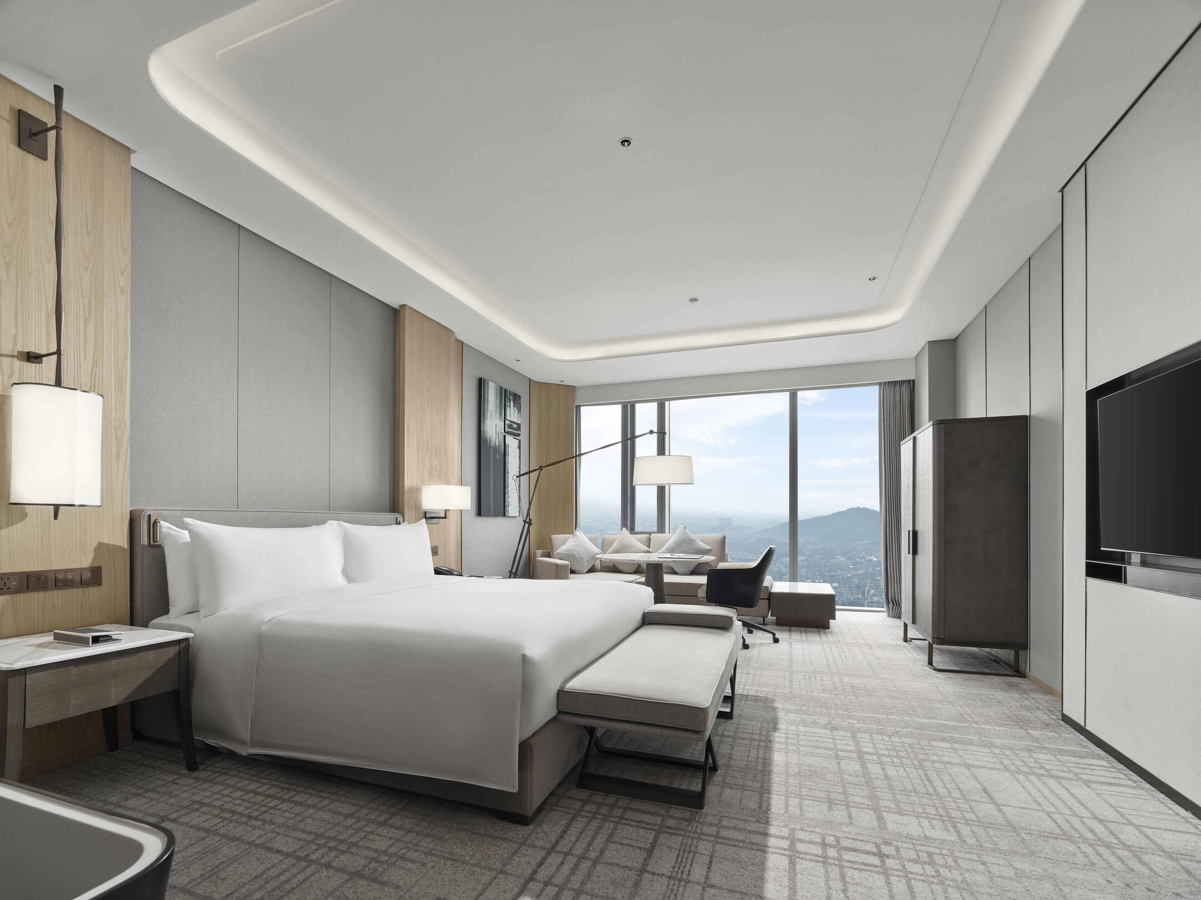 A modern hotel room with a large bed, a desk, a chair, a TV, and a view of the city.