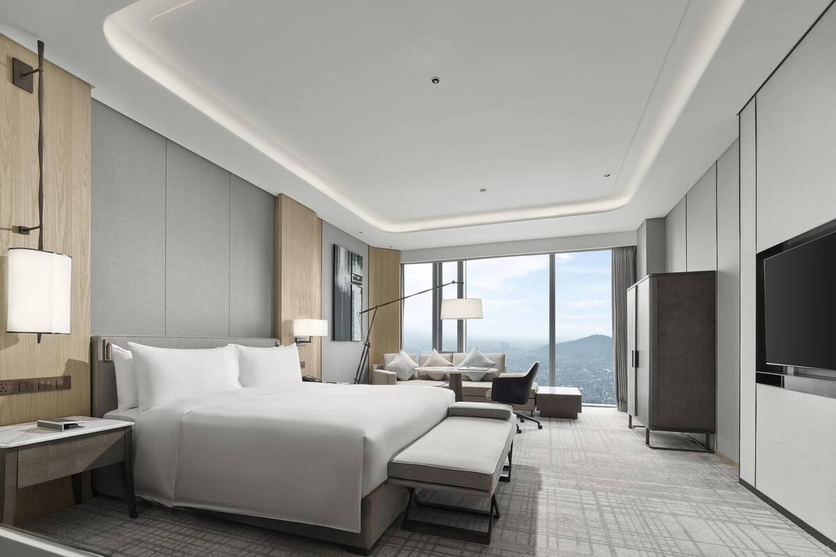 A modern hotel room with a large bed, a desk, a chair, a TV, and a view of the city.
