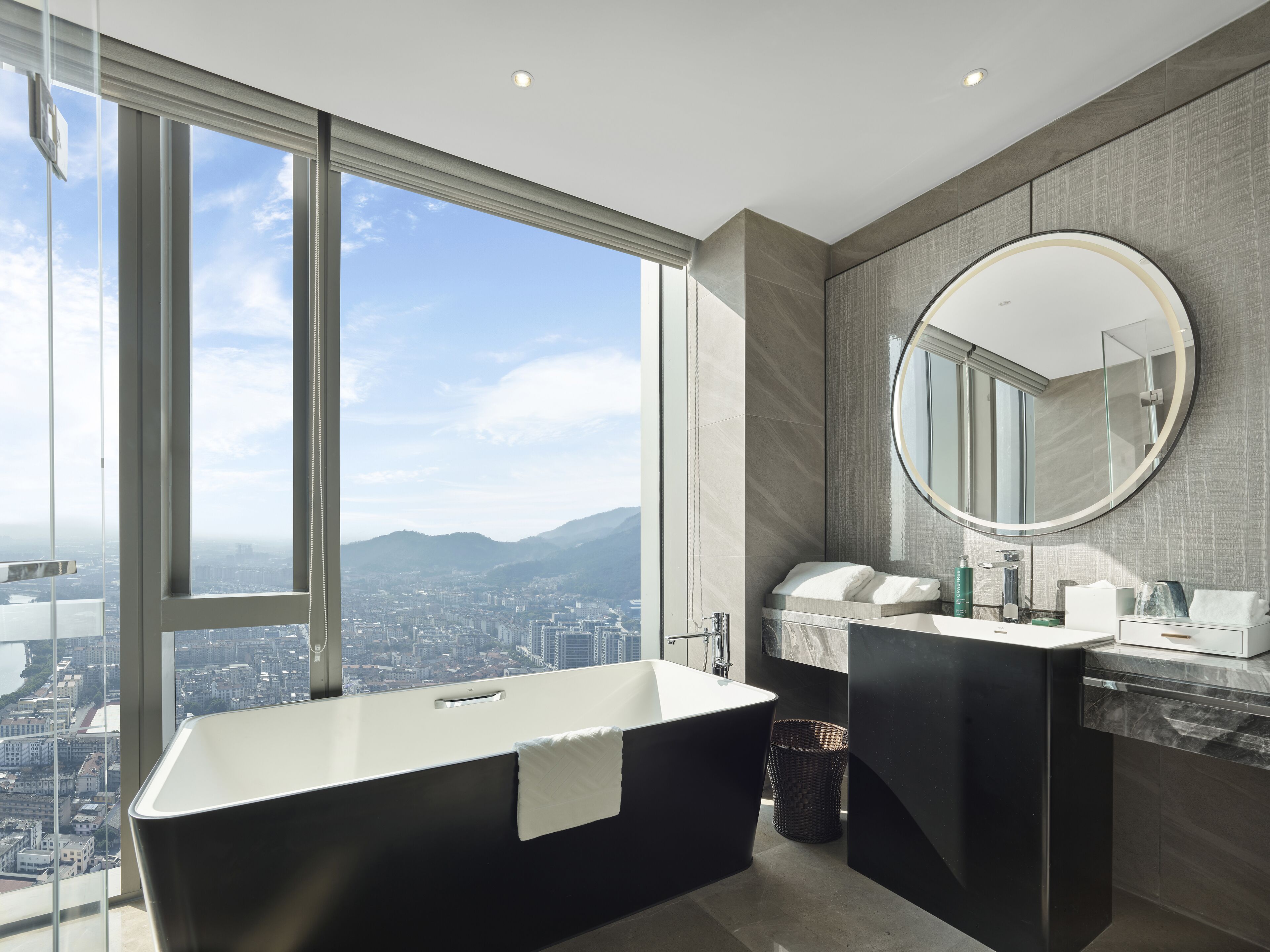Une salle de bain moderne avec une grande baignoire, un grand miroir rond et une vue sur la ville grâce à de larges fenêtres.
