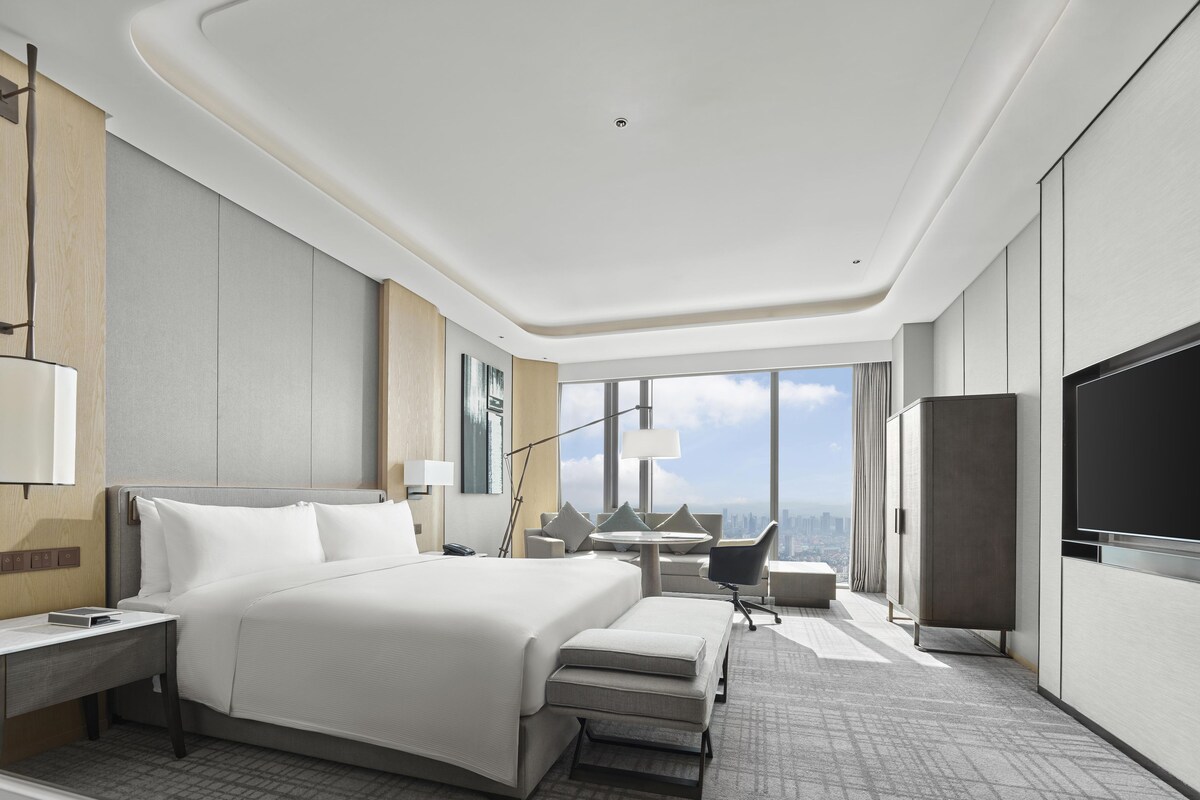 A modern hotel room with a large bed, a desk, a chair, a TV, and a city view.
