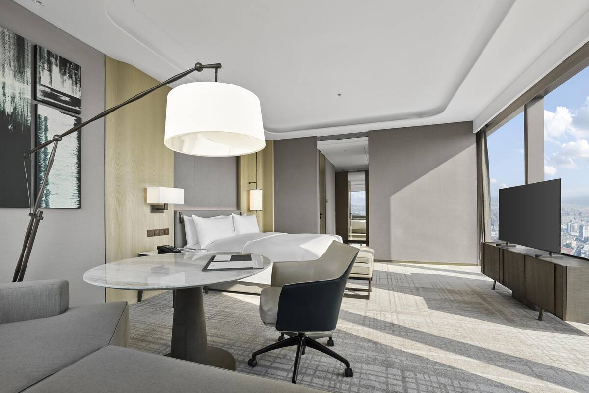 A modern hotel room with a bed, a desk, a chair, a TV, and a large window offering a city view.