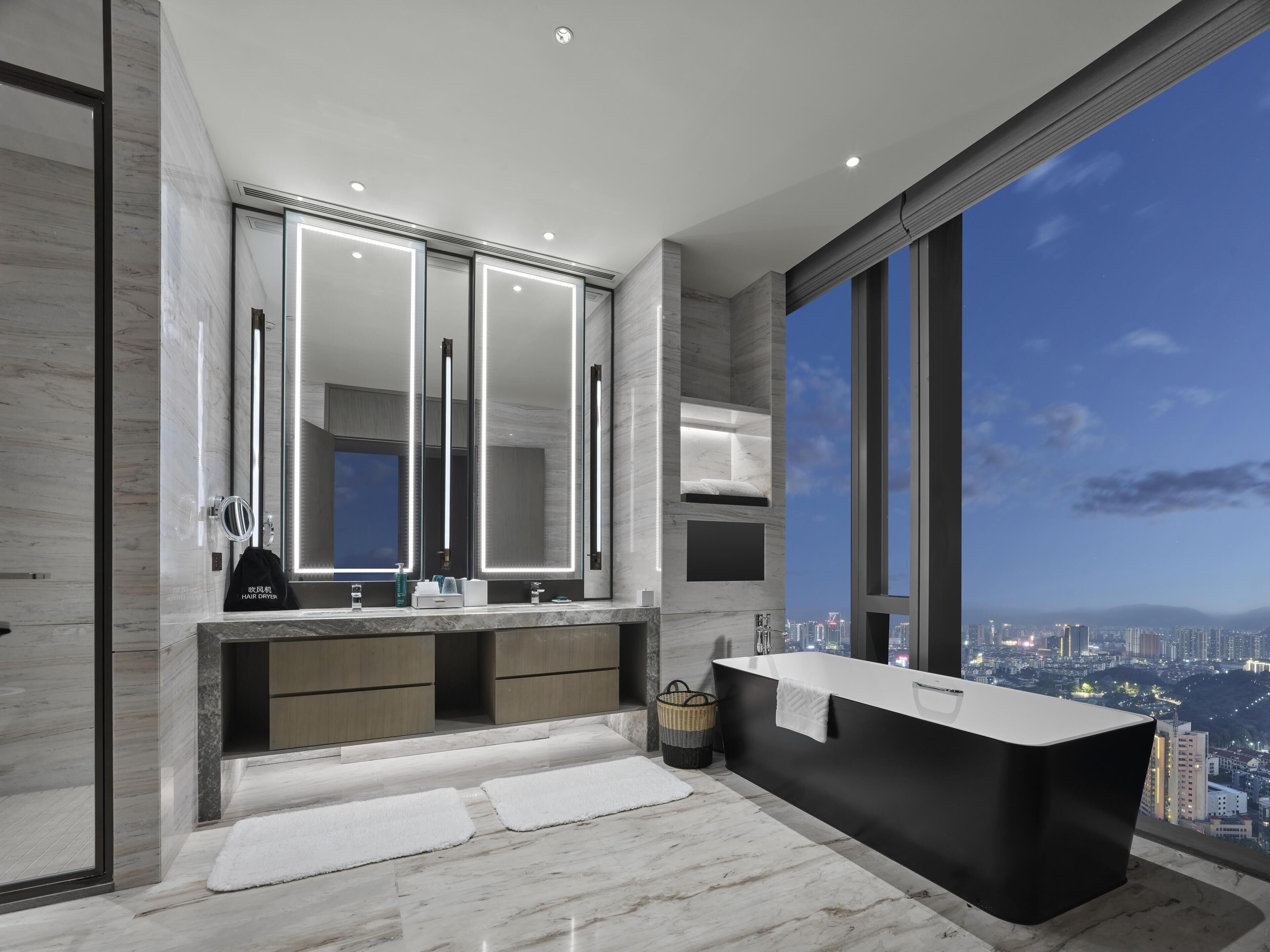 Un baño moderno con un espejo grande, lavamanos dobles, una bañera y vistas a la ciudad.