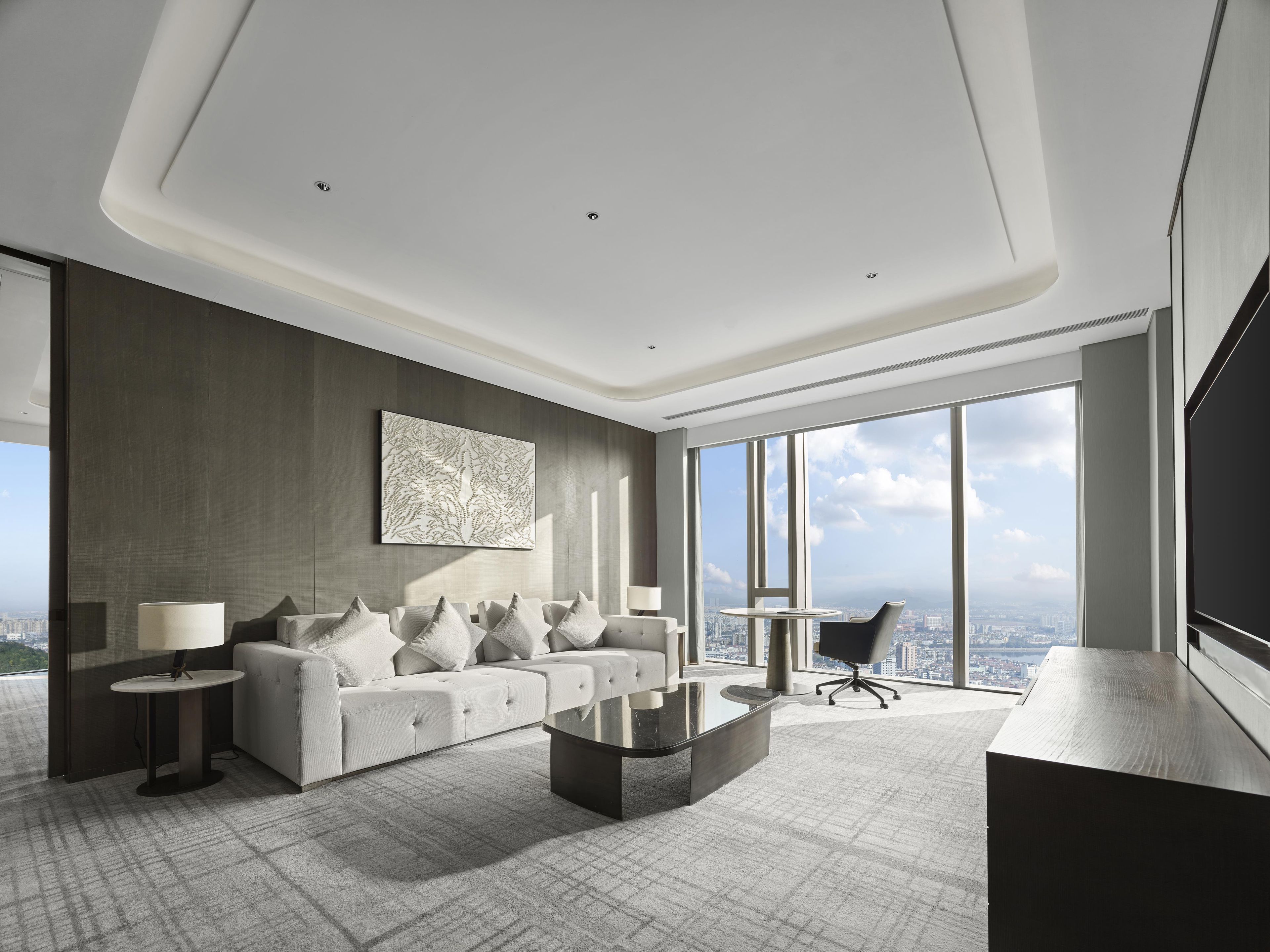 A modern hotel room with a large window, a sofa set, a coffee table, and a desk.