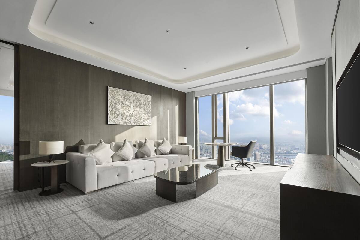 A modern hotel room with a large window, a sofa, a coffee table, and a desk.
