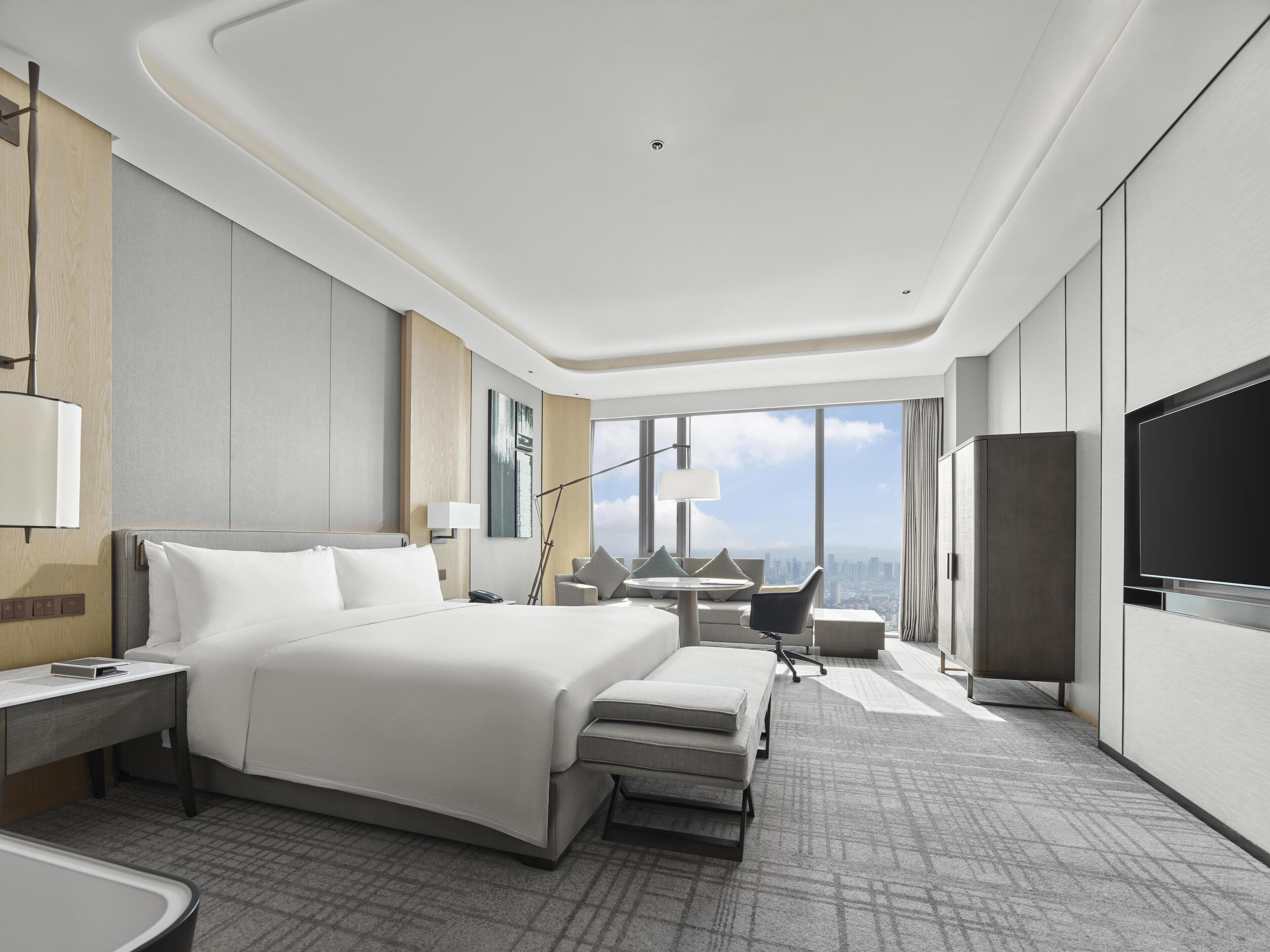 A modern hotel room with a large bed, a desk, a chair, a TV, and a city view.