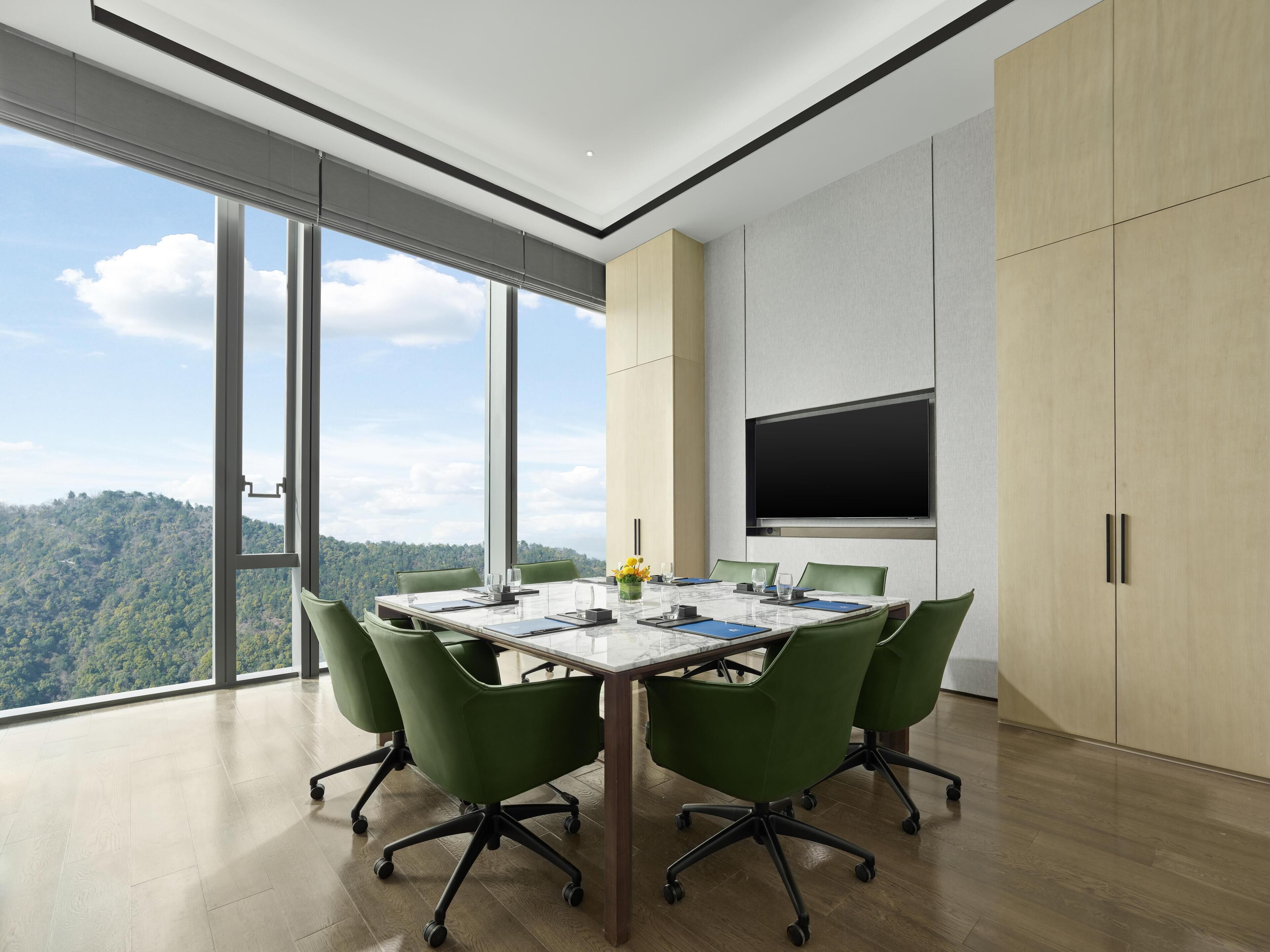 Un salón de conferencias con una gran mesa, sillas verdes, un televisor de pantalla plana y vistas a una colina boscosa.