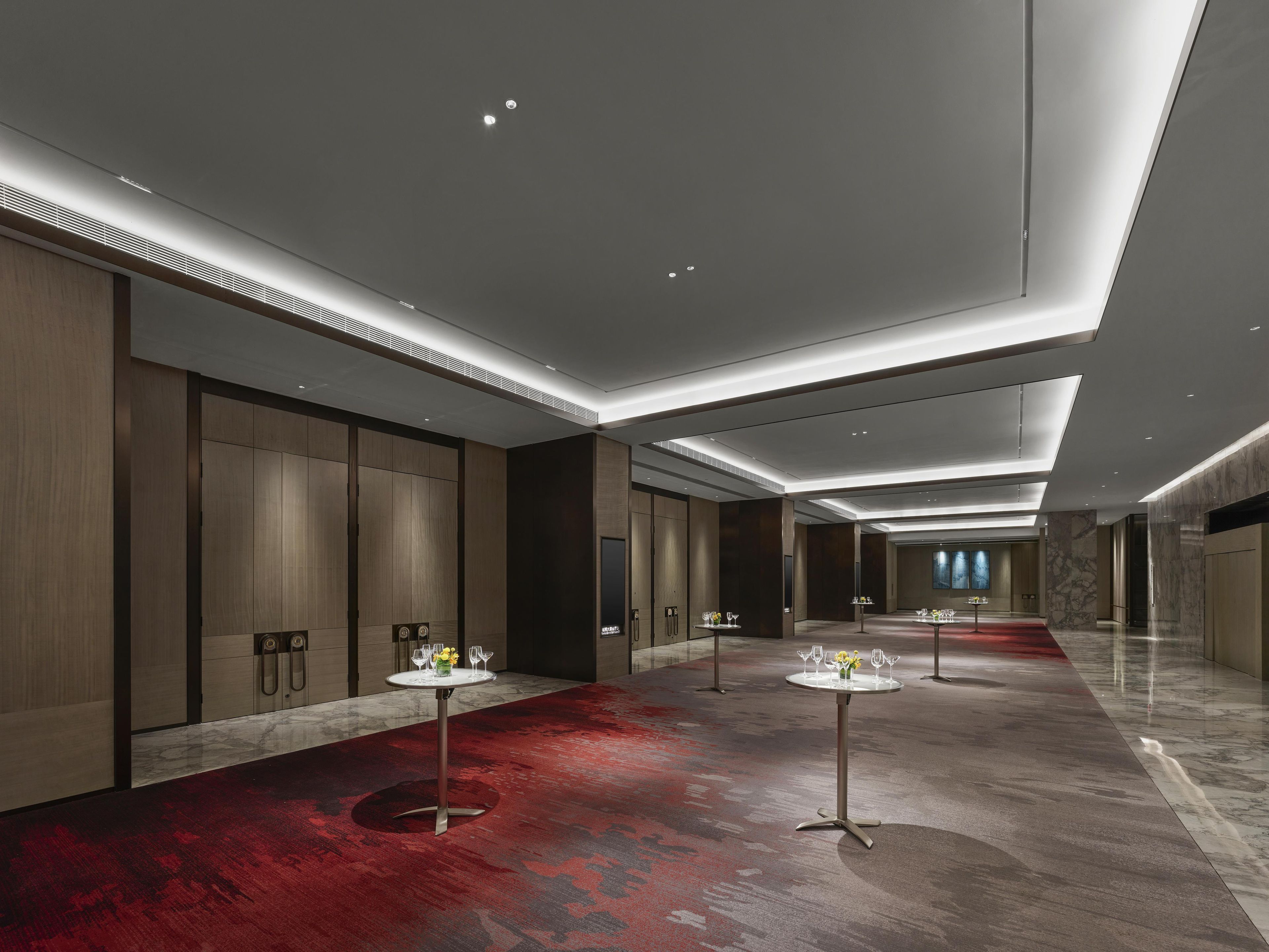 A modern hotel corridor with a red carpet, wooden paneling, and a long reception desk with a vase of flowers.