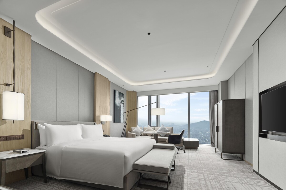 A modern hotel room with a large bed, a desk, a chair, a TV, and a view of the city.