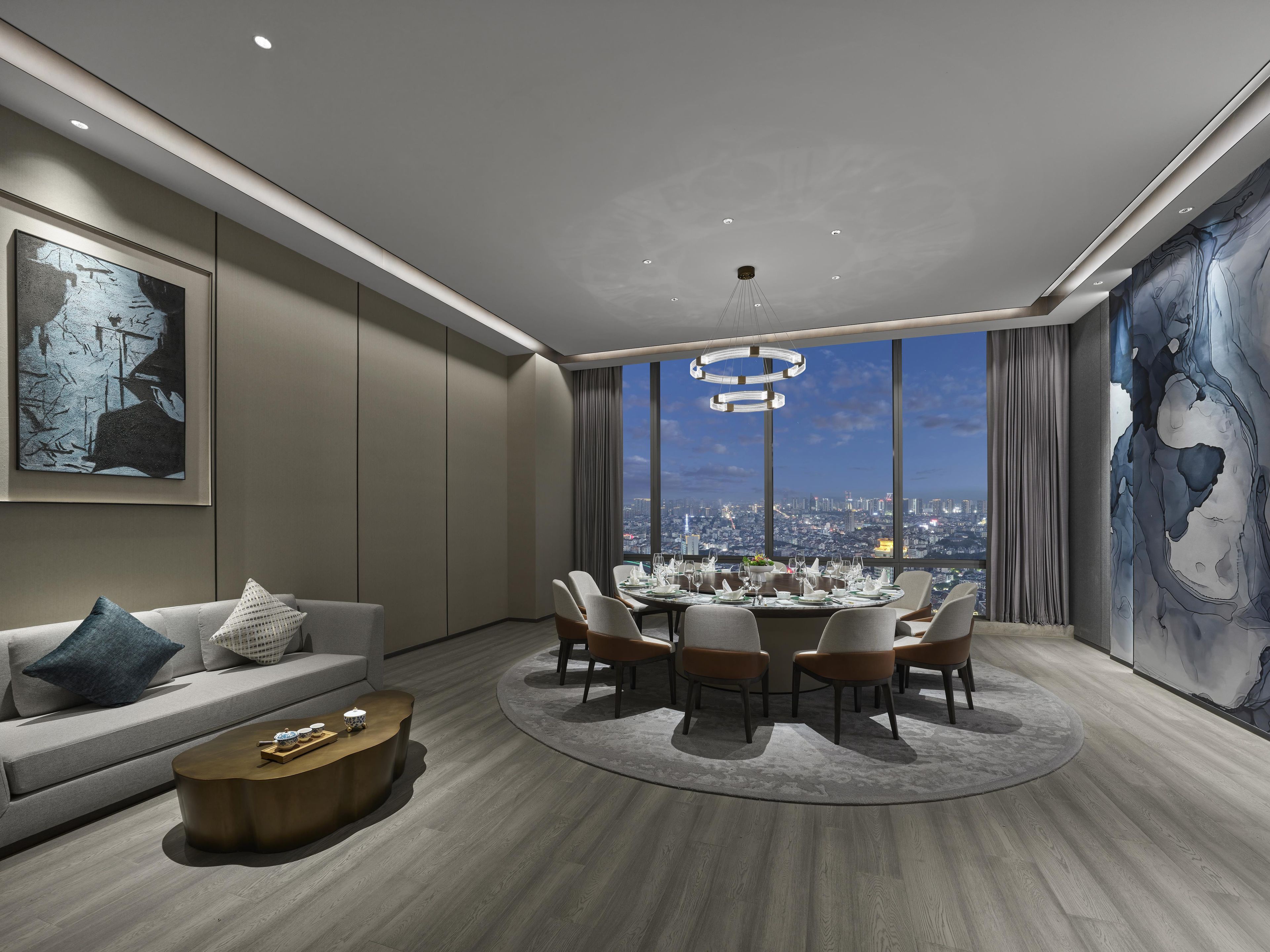 A modern hotel room with a dining area, a sofa, and a large window offering a city view.