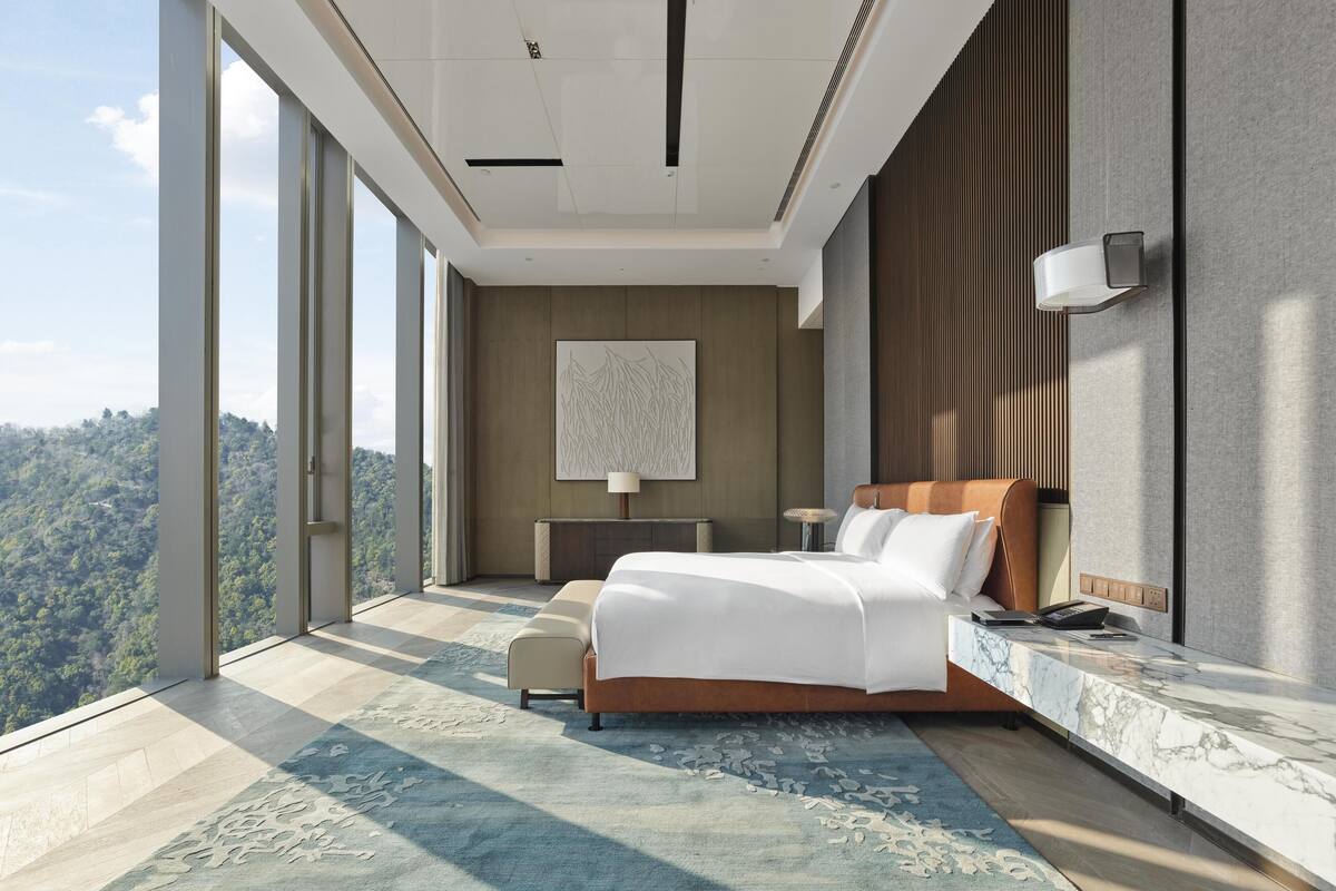 A modern hotel room with a large bed, a desk, and a view of a forested area.