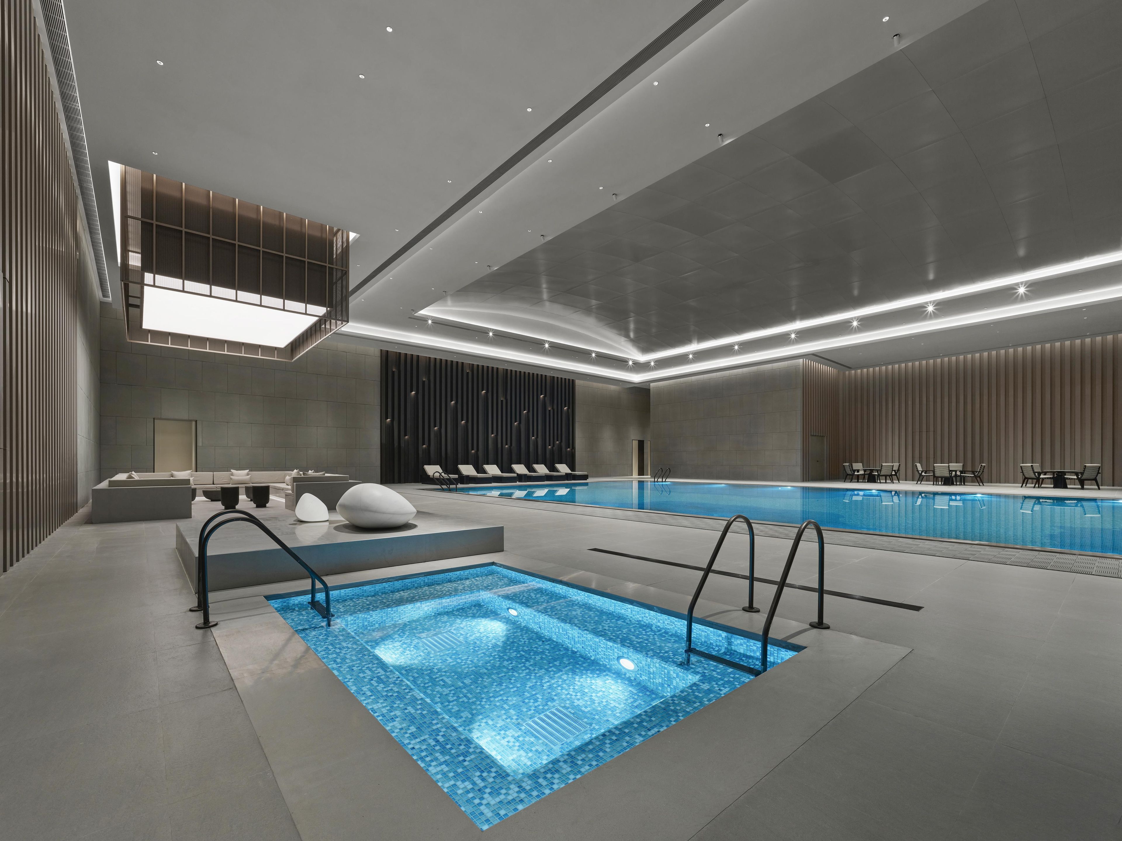 Piscina interior moderna de forma rectangular, rodeada de sillones de descanso y una zona de estar con bar junto a la piscina.