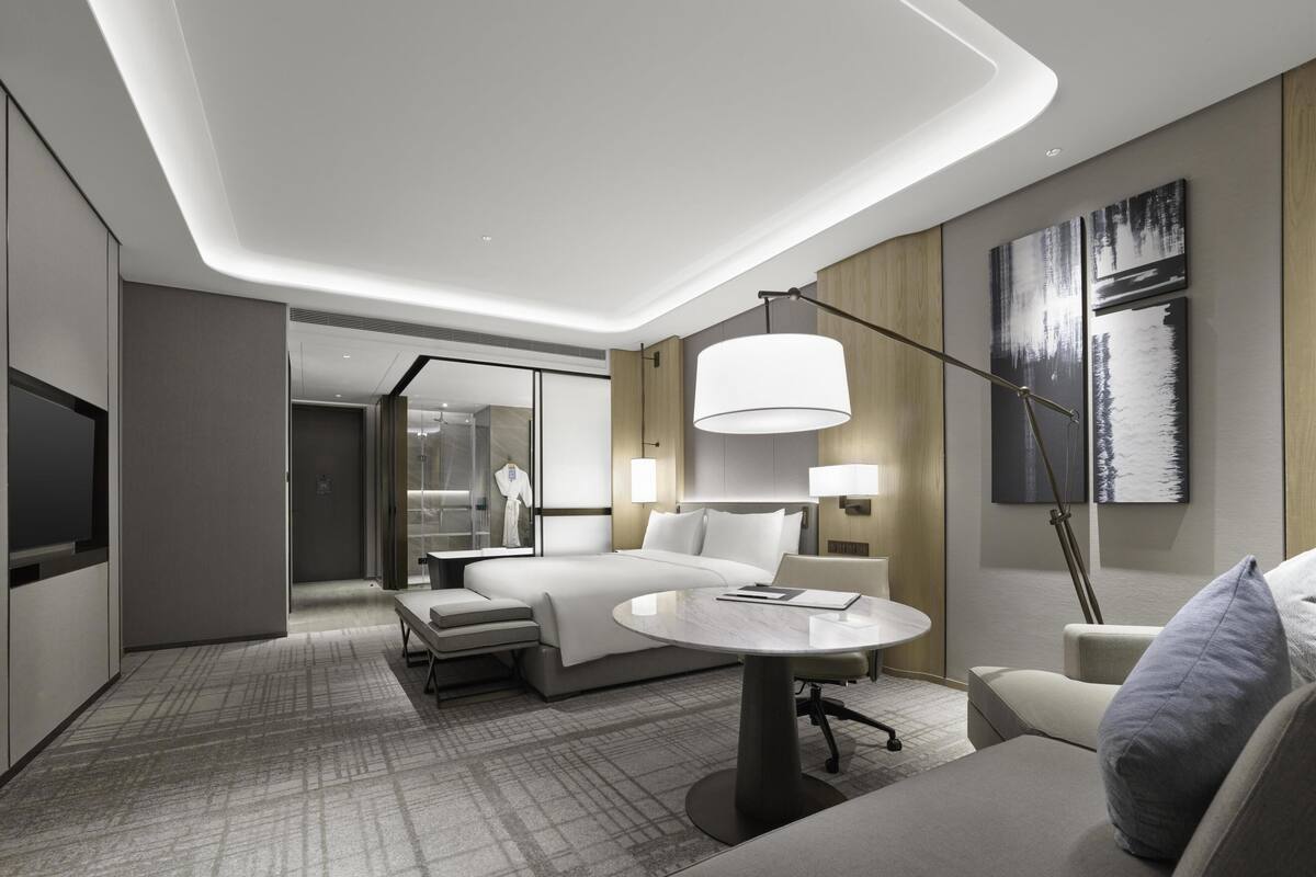 A modern hotel room with a bed, bedside tables, a round table, a sofa, and a TV.