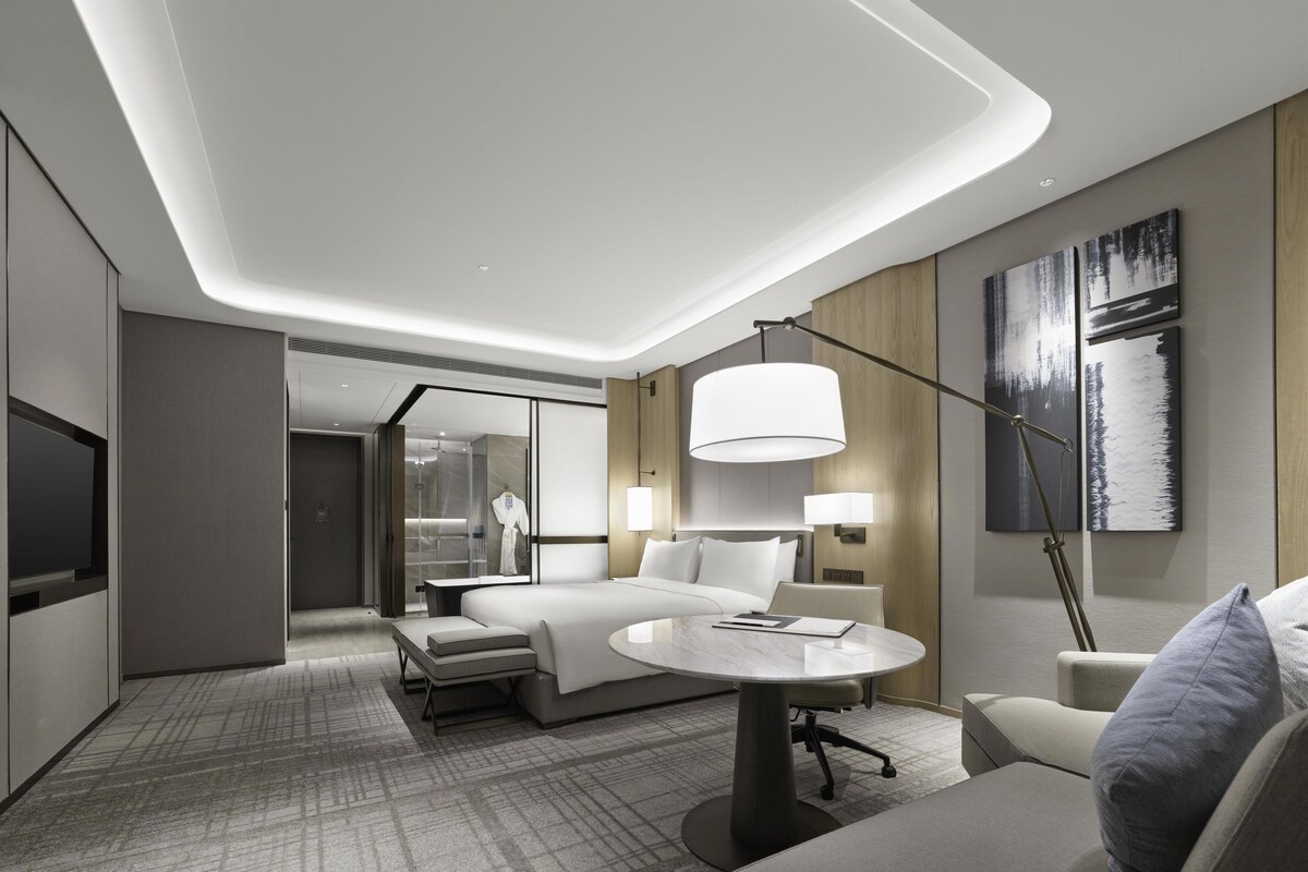 A modern hotel room with a bed, bedside tables, a desk, a chair, a TV, and artwork on the walls.
