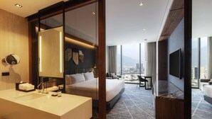 A modern hotel room with a large bed, a separate sitting area, a flat-screen TV, a large window with a city view, and a bathroom with a sink and mirror.