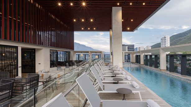 A rooftop pool area with lounge chairs and tables, a glass railing, and a view of mountains.