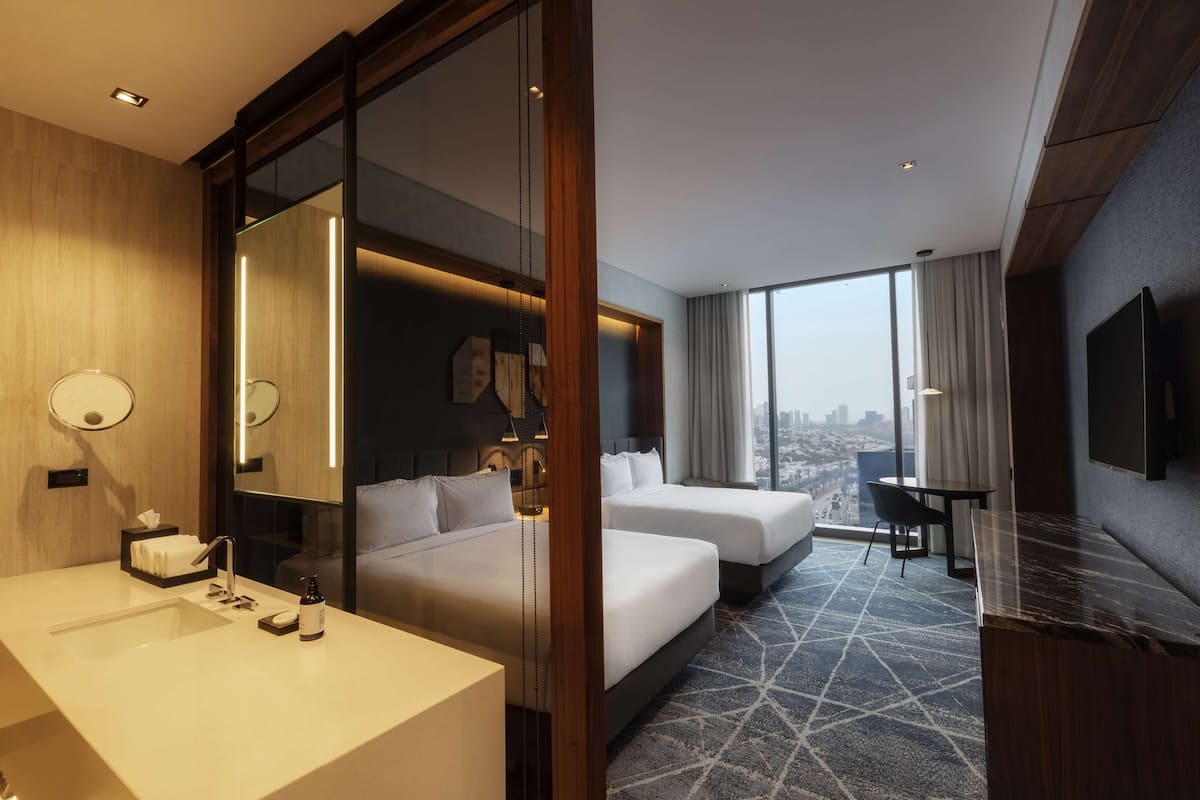 A hotel room with a large bed, a desk, a chair, a TV, a bathroom with a sink and mirror, and a city view.