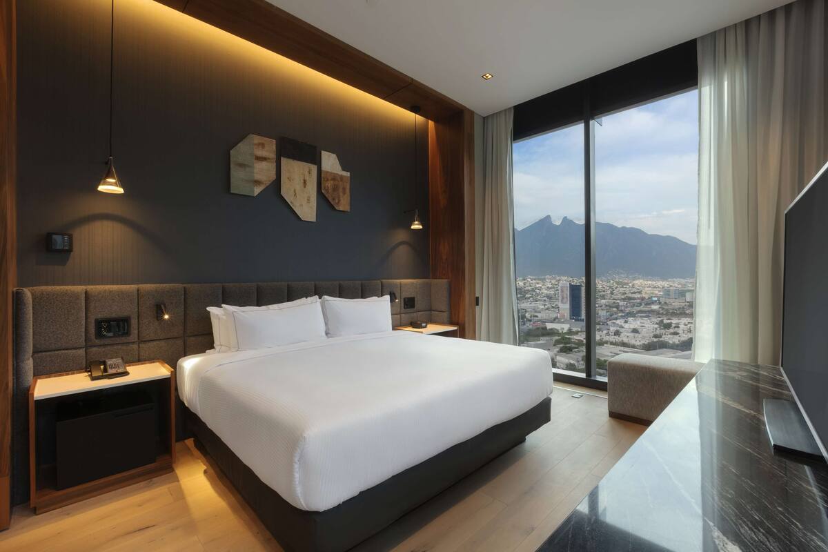 A modern hotel room with a large bed, bedside tables, a nightstand, and a view of a cityscape through large windows.